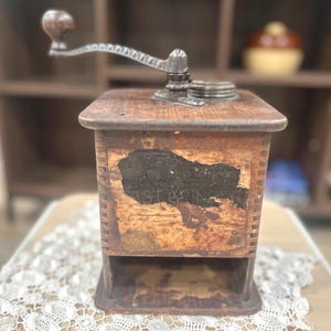 May include: Antique wooden coffee grinder with a metal crank handle. The grinder has a rectangular wooden body with a faded label and a small drawer at the bottom. The wood has a distressed, aged appearance, suggesting it is vintage.