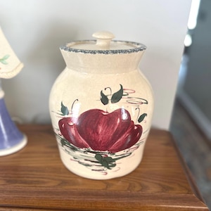 May include: Cream-colored ceramic jar with a lid, featuring a hand-painted design of red apples with green leaves. The jar has a blue-grey rim and sits on a wooden surface. A decorative kitchen storage container.