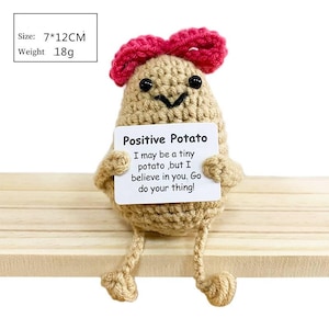 May include: A crocheted potato figure with a red bow, black eyes, and a smiling mouth. It holds a sign that reads "Positive Potato" with an inspirational message. The figure is approximately 2.75 x 4.75 inches.