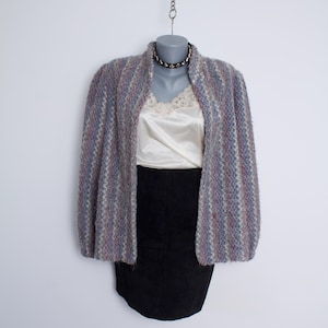 May include: A multi-colored knit cardigan with shades of purple, blue, and gray, displayed on a mannequin. The cardigan is paired with a cream-colored camisole and a black skirt. A black choker necklace is also visible.