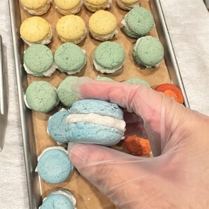 以下が含まれることがあります： 青、緑、黄色など、白いフィリングが入ったカラフルなマカロンのトレイ。青いマカロンが手袋をはめた手で持たれています。マカロンはベーキングシートに並べられています。