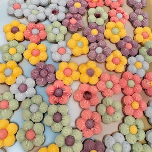 May include: A selection of flower-shaped sweets in a variety of colours. The sweets are in shades of yellow, pink, purple, green, and white, each with a contrasting centre. The sweets are arranged closely together, forming an attractive pattern.