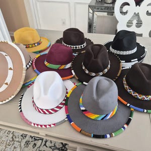 May include: A collection of fedora hats in various colors, including brown, yellow, white, and gray. Each hat features a beaded band around the crown and brim, with colorful patterns. The hats are arranged on a light-colored surface.
