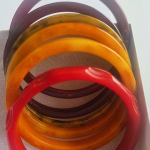May include: A collection of colorful bangles in a variety of shades, including red, orange, yellow, and deep purple. The bracelets are stacked together, showcasing their smooth, glossy surfaces. The bangles are likely made of a hard plastic or resin material.