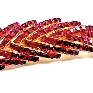 May include: A gold-tone metal brooch shaped like a curved feather or leaf, featuring multiple rows of faceted, deep magenta-colored crystals arranged in a chevron pattern along the length of the piece, creating a textured and reflective surface.