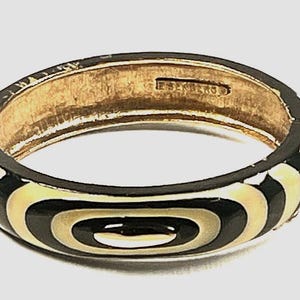 May include: A gold-toned hinged bangle bracelet with a black and cream-colored geometric design. The bracelet features a series of concentric oval shapes in black and cream. The interior of the bracelet is gold-toned, and the clasp is visible.