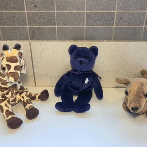 May include: Three plush toys: a giraffe with brown spots, a navy blue bear with a ribbon, and a tan dog with floppy ears. The giraffe and dog are lying down, while the bear is sitting upright.