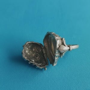Puede incluir: Un anillo de metal plateado con una parte superior articulada tipo relicario que se abre para revelar un interior hueco. La superficie exterior del relicario tiene un acabado orgánico texturizado y la banda tiene un detalle metálico decorativo.