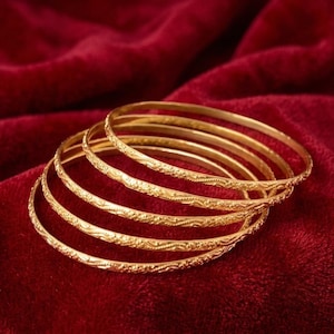 Pode incluir: Um conjunto de cinco pulseiras finas de ouro, cada uma com um padrão delicado e repetitivo em relevo na superfície externa. As pulseiras circulares estão empilhadas, exibindo um acabamento metálico polido que reflete a luz nas bandas.
