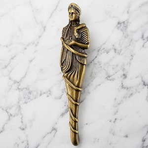 May include: A vertical brass-toned metal door handle shaped like a standing figure wearing a draped garment. The figure has a braided hairstyle and a snake coiled around the torso and lower body, which tapers into a smooth, elongated base.