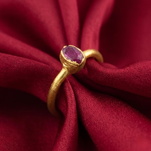 May include: A gold ring with a ruby gemstone. The ring has a textured band and a bezel setting holding the oval, deep red ruby. The ring is displayed on red satin fabric.