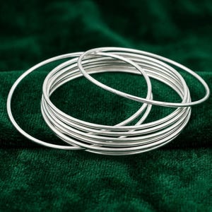 Könnte beinhalten: Ein Set aus mehreren dünnen, polierten silberfarbenen Armreifen. Die kreisförmigen Metallbänder sind glatt und reflektierend und weisen eine einfache, runde Drahtkonstruktion auf. Die Armreifen sind gestapelt und erzeugen eine geschichtete Optik.