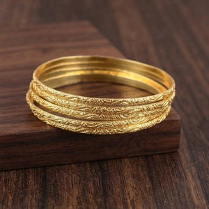 Pode incluir: Um conjunto de três pulseiras rígidas de cor dourada com um padrão em relevo contínuo e intrincado em toda a superfície. Cada pulseira circular tem um acabamento polido e uma largura consistente, criando um visual empilhado.