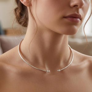 Op de afbeelding: Een zilverkleurige choker ketting. De ketting heeft een eenvoudig, gebogen ontwerp dat dicht op de nek zit. De uiteinden van de ketting hebben kleine, ronde, decoratieve accenten. De ketting wordt door een persoon gedragen.