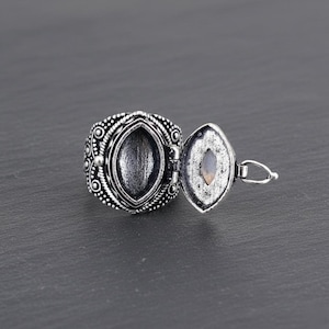 Peut inclure: Bague locket argentée avec un compartiment ovale qui s'ouvre pour révéler une petite pierre. La bague présente un motif orné et texturé autour de l'anneau et du médaillon. Le fond est gris foncé.