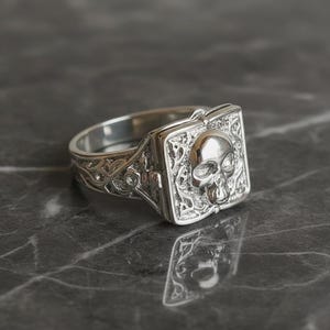 May include: A silver-toned ring featuring a square face with a raised, polished skull emblem centered on a textured, intricate background. The band of the ring displays detailed, ornate scrollwork patterns that extend from the sides to the shank.