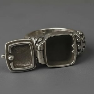 Peut inclure: Une bague poison en métal argenté dotée d'un compartiment carré à charnière qui s'ouvre pour révéler un intérieur creux. L'anneau est orné d'un motif perlé texturé sur les côtés, et le couvercle comprend un petit bouton sphérique.