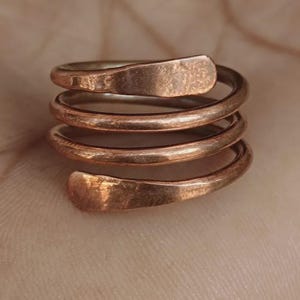 Puede incluir: Un anillo de metal color cobre con un diseño en espiral de tres vueltas. La superficie del metal presenta un acabado martillado y texturizado, y los extremos del alambre están aplanados y redondeados, creando una apariencia envolvente.