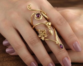 May include: A gold-toned multi-finger ring featuring a swirling wire design, a small five-petaled flower, and two round purple gemstones. The metal structure extends across the finger with delicate leaf accents and a polished finish.