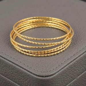 May include: A set of five thin gold bangles, each featuring a delicate, repeating textured pattern along the entire circumference. The bracelets are stacked together, showcasing a bright, metallic yellow finish and a consistent, slender circular design.
