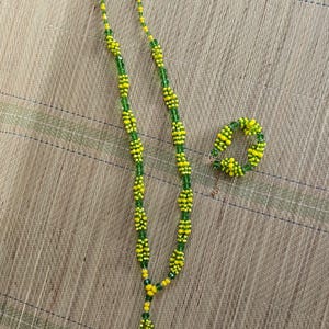 Può includere: Un set di collana e braccialetto di perline. La collana, con nappina, e il braccialetto, un semplice anello, sono realizzati con perline gialle e verdi. Il design è vivace e accattivante. Su un tappeto intrecciato.
