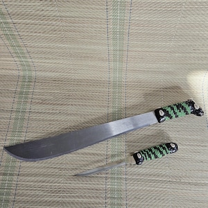 May include: A large, silver machete and a smaller knife with black and green beaded handles. The blades are silver and the handles are black with green accents. The knives are displayed on a woven mat.