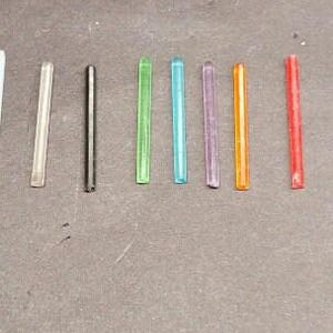May include: A selection of coloured glass rods. The rods are arranged horizontally and vary in colour, including clear, black, green, blue, purple, orange, and red. They are all approximately the same length.
