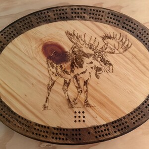May include: An oval wooden cribbage board featuring a dark brown outer track with drilled peg holes and a lighter wood center. A detailed wood-burned illustration of a moose stands in the middle, positioned near a prominent dark wood knot.