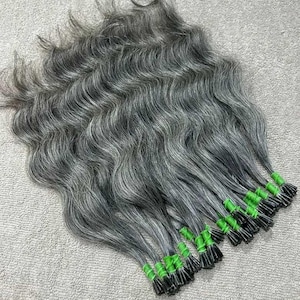 May include: A collection of wavy, grey hair extensions with black tips. The hair is tied in small bundles with green bands. The extensions are arranged on a light grey surface.