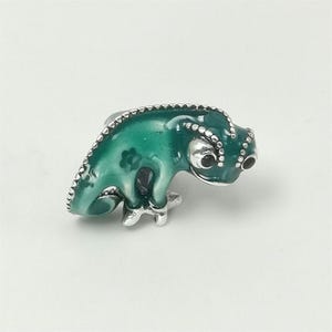 May include: A green and silver chameleon ring. The ring features a detailed, enameled chameleon with a textured body and silver accents. The chameleon is in a side-facing pose, with its eyes and feet clearly visible. The ring is set against a plain white background.