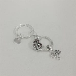 Puede incluir: Un llavero de metal plateado con una anilla circular y una pequeña etiqueta redonda, unido a un soporte circular texturizado. Incluye un dije en forma de corazón con piedras rojas y un dije colgante de corazón con filigrana calada.