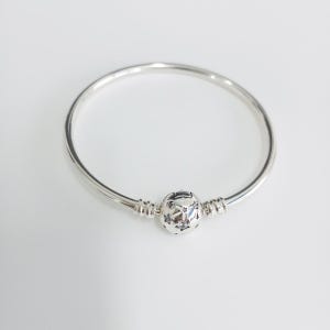 May include: A silver bangle bracelet with a decorative clasp. The clasp has a detailed design, and the bracelet has a smooth, polished finish. The bracelet is a simple, elegant piece of jewellery.