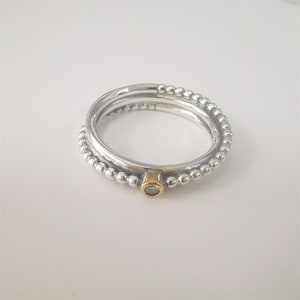 Puede incluir: Un conjunto de tres anillos de plata. Dos anillos tienen un diseño de cuentas y el tercero es liso. Un anillo presenta un pequeño acento dorado con una piedra transparente. Los anillos están apilados.