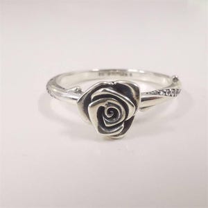 Op de afbeelding: Een zilveren ring met een gedetailleerd rozenontwerp. De roos is het middelpunt, met ingewikkelde details van de bloemblaadjes. De band heeft kleine, ingebedde stenen aan één kant. De ring staat tegen een effen witte achtergrond.