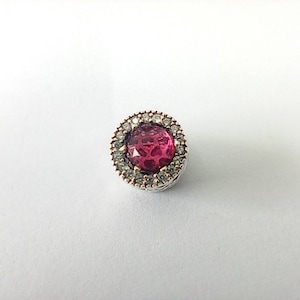 May include: A round, silver charm with a large, faceted, magenta-colored center stone. The stone is surrounded by a halo of smaller, clear, sparkling stones. The charm is set against a white background.