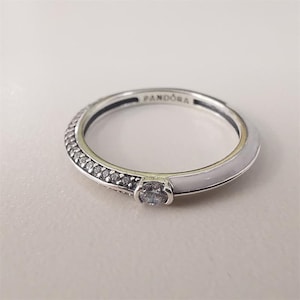 Könnte beinhalten: Ein silberner Ring mit einem weißen Emailleband und einer Reihe kleiner, klarer Steine. Der Ring hat einen einzelnen, größeren klaren Stein und das Wort "PANDORA" ist auf dem Band eingraviert.