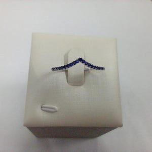 Può includere: Un anello color argento con un design a forma di V, impreziosito da piccole pietre preziose blu scuro. L'anello è esposto su un supporto bianco strutturato.