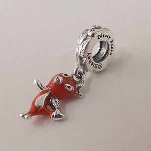 Op de afbeelding: Een zilveren bedel met een feloranje geëmailleerd figuurtje dat aan een rond oogje hangt. Het oogje is gegraveerd met de tekst Disney Parks en heeft een gestructureerd, bladachtig patroon langs de buitenrand.