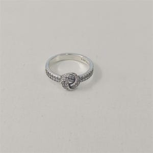 Può includere: Un anello d'argento con un design a nodo. L'anello presenta una fascia con piccole pietre trasparenti e un elemento centrale a forma di nodo anch'esso ornato da pietre trasparenti. L'anello è posto su uno sfondo bianco.