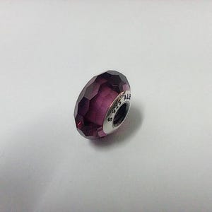 Puede incluir: Una cuenta de dije facetada de color púrpura intenso con una banda plateada. La cuenta tiene un diseño geométrico y está marcada con "S 925 ALE". Este artículo de joyería es adecuado para pulseras y collares.