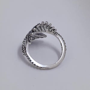 May include: A silver ring with a leaf design. The ring features intricate detailing, including small dots and lines, creating a textured appearance. The ring is set against a plain white background.