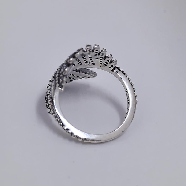 May include: A silver ring with a leaf design. The ring features intricate detailing, including small dots and lines, creating a textured appearance. The ring is set against a plain white background.