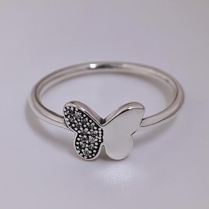 May include: A silver ring with a butterfly motif. One wing of the butterfly is embellished with small, dark gemstones, the other is plain. The ring has a slender band and a polished finish, suitable for everyday wear.