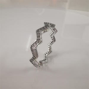 Puede incluir: Un anillo plateado con un diseño en zigzag. El anillo está incrustado con pequeñas piedras oscuras a lo largo de los bordes y tiene las letras "ALE". El anillo está sobre una superficie blanca.