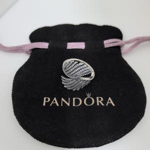 May include: A silver ring with a feather design, encrusted with small, clear gemstones. The ring sits on a black velvet pouch with the brand name "PANDORA" printed in white. The pouch has a pink drawstring.