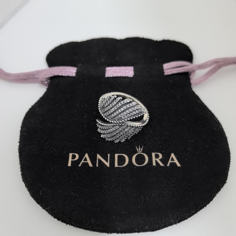 May include: A silver ring with a feather design, encrusted with small, clear gemstones. The ring sits on a black velvet pouch with the brand name "PANDORA" printed in white. The pouch has a pink drawstring.