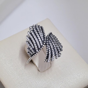 May include: A silver-toned ring in the shape of two stylized wings. Each wing is covered in small, sparkling clear stones. The ring is set against a neutral background, highlighting its intricate design and details.