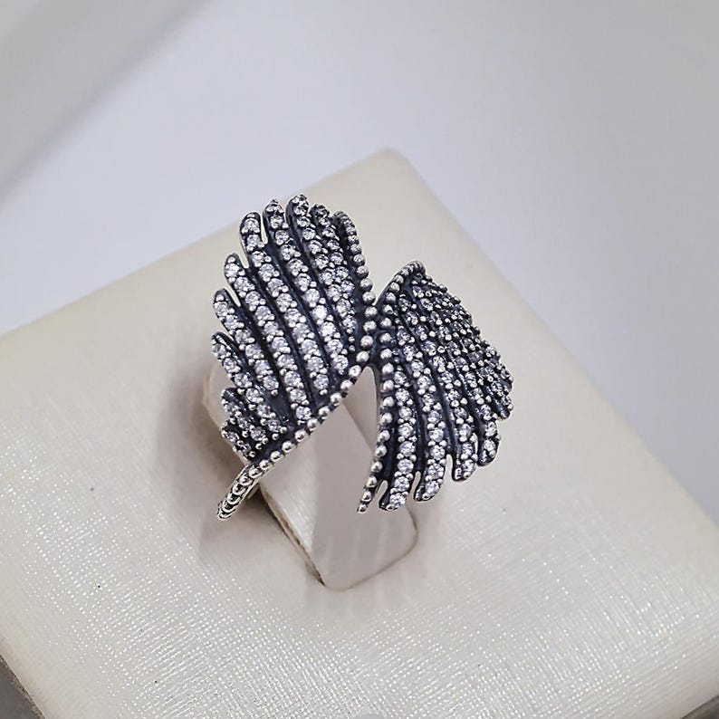 May include: A silver-toned ring in the shape of two stylized wings. Each wing is covered in small, sparkling clear stones. The ring is set against a neutral background, highlighting its intricate design and details.