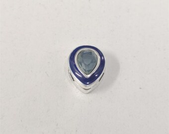 Op de afbeelding: Een zilveren bedel met een druppelvormige, lichtblauwe edelsteen. De edelsteen is omgeven door een donkerblauwe emaille rand. De bedel heeft een gepolijste afwerking.