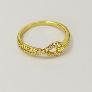 May include: A gold-colored ring with a unique infinity symbol design. The ring features a band of small, clear stones set in a curved pattern, creating a sparkling effect. The ring is a piece of jewelry.
